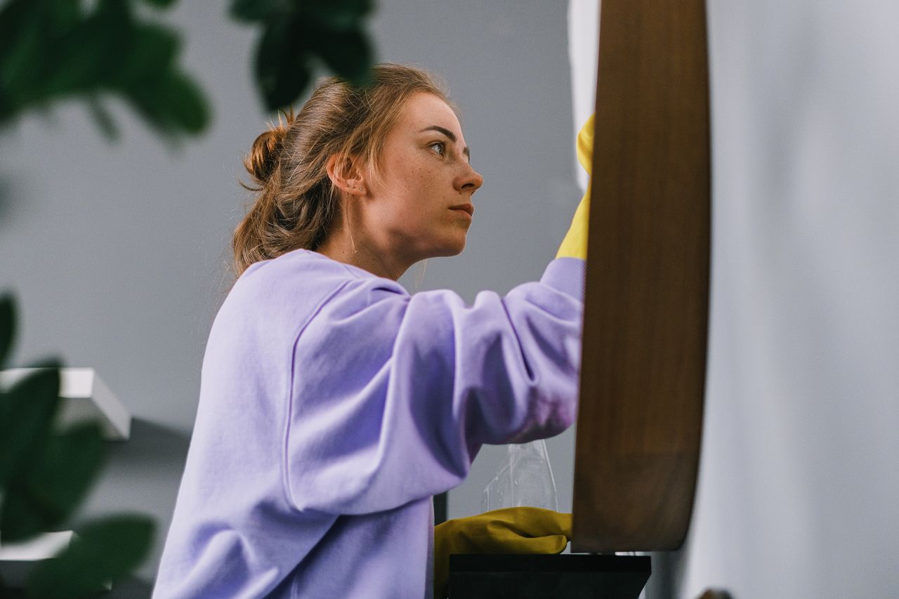 Side view of a young focused woman in latex gloves standing and cleaning a mirror in a bright room during the day